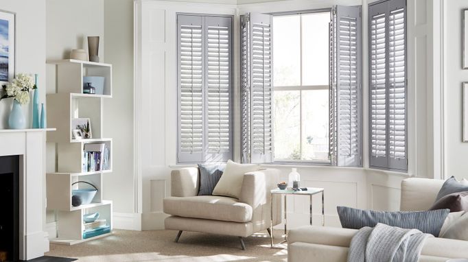 Grey full height shutters from housebeautiful range dressed on windows of cream living room. Cream sofa chairs can also be seen in the view.