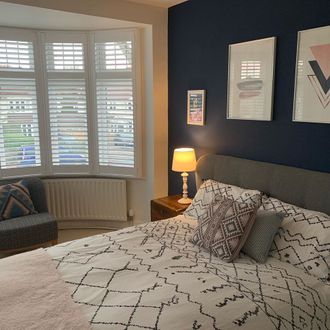 Bedroom shot, white patterned sheets, pink throw and dark blue walls featuring bay window white shutters