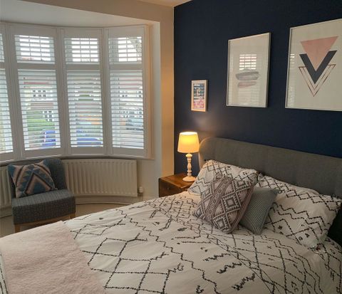 Bedroom shot, white patterned sheets, pink throw and dark blue walls featuring bay window white shutters