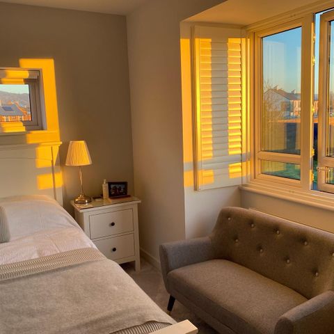 White shutters in bedroom behind grey sofa seat with sunset coming through windows