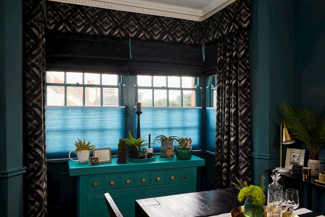 Black roman blinds and teal pleatedblinds are fitted to the same rectangular windows along with gasoline decorated curtains in a dining team decorated with dark and blue colours