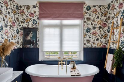 A red roman blind paired with a white transition blind on a window in a bathroom with floral wallpaper, blue walls and a bathtub in the middle of the room