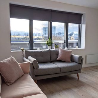 Acacia Gunmetal Roller blind on window with view of city behind grey sofa with pink cushion and pink sofa