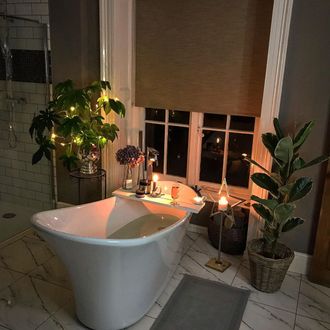 Beige roller blind in bathroom at night in front of shaped bath and plants, candles and prosecco glass