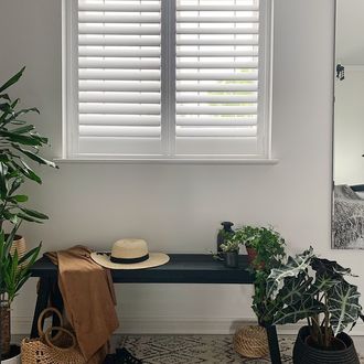 White shutters on white wall with black bench, straw hat, bag, sandals and plants.