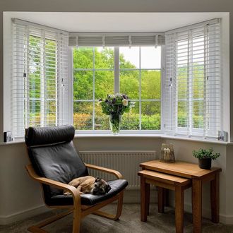 white Venetian blinds in bay window with leather arm chair with dog asleep on and vase of flowers in the windowsill.
