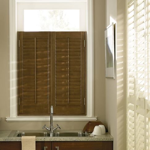 Dark brown coloured shutters fitted above a sink to a tall rectangular window in a kitchen decorated with cream walls