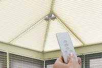 hand holding a remote infront of four motorised cream pleated blinds on a conservatory roof 