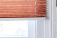 close up of orange pleated blind with fabric overhang in front of white window 