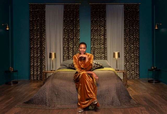 someone sat on the edge of a bed in an orange velvet robe in a room with wooden floors, teal blue walls and curtains matched with white voile curtains