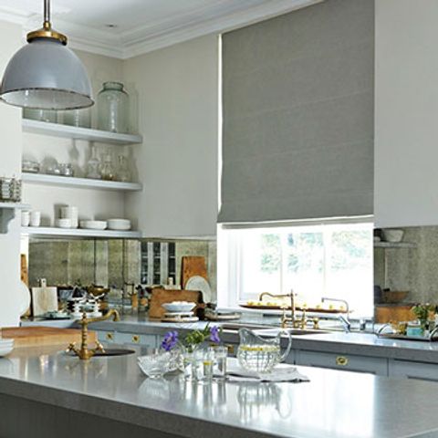 Grey coloured roman blind is fitted to a tall rectangular window in a kitchen with grey countertops, white walls and a range of kitchenware