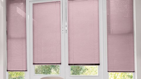 French doors which are fitted with Portfolio Pink Blossom in a Pink tone. The Perfect Fit venetian blinds are a quarter open, allowing small amounts of light to shine onto the white dining room chairs and cream topped dining room table.