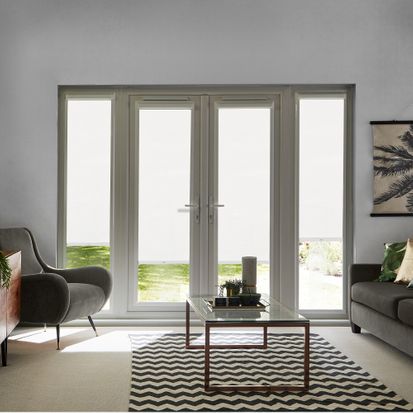 French doors which are fitted with Jerico Snow in a white tone. The Perfect Fit Roller blinds are half-open, allowing small amounts of light to shine onto the armchair, coffee table and sofa that are next to the doors.