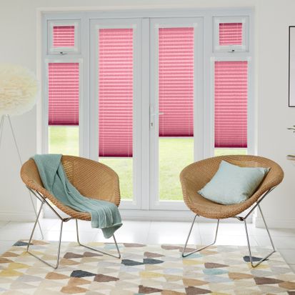 French doors which are fitted with Lanbury Blackout Pink in a Pink tone. The Perfect Fit Pleated blinds are a quarter open, allowing small amounts of light to shine onto the white walls and neutral wicker chairs.