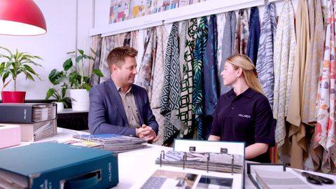 George Clarke sits with female advisor with vibrant fabrics hanging behind them and books of fabric swatches on the table