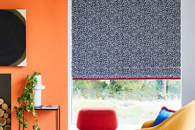 Blue and white patterned roman curtain fitted to a tall rectangular window in a room with orange walls, yellow chair and red pouffe stool
