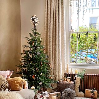 Christmas tree sat infront of a window in a neutral living room with neutral curtains draped to the floor