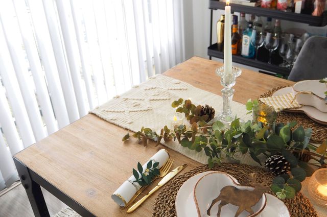 Christmas dining table place set with gold coloured cutlery, candles and foliage, white vertical blinds in the background