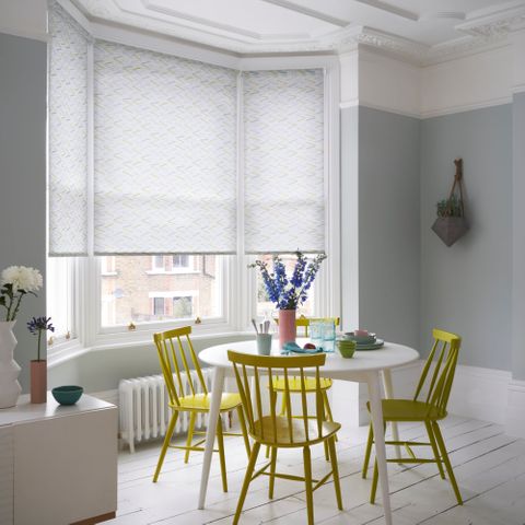 white roller blinds with bright details in a bay window in a dining room with funky bright yellow dining chairs