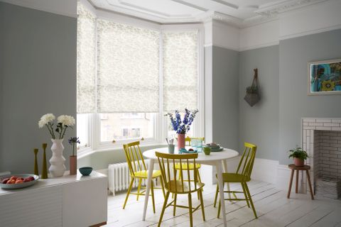 A neutral blue dining room with white and muted yellow, floral, bay-window blinds and striking yellow furniture