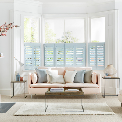 light blue cafe style shutters fitted to a bay window in a living room decorated in white with a rose pink sofa, armchair and a glass coffee table 