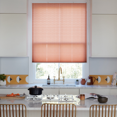pink coloured pleated blinds fitted to a tall rectangular window in a kitchen decorated entirely in white with copper coloured chairs and tap