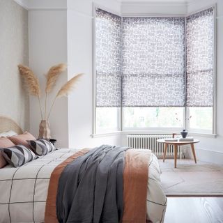Roller blinds for a bay window in a bedroom with a bed on the left side of the room and a small table and radiator by the window.