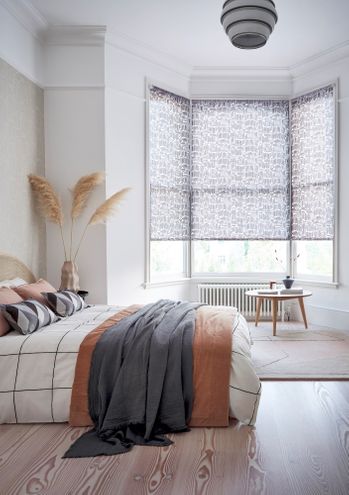 Roller blinds for a bay window in a bedroom with a bed on the left side of the room and a small table and radiator by the window.