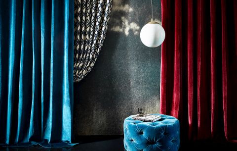 blue and red velvet curtains against a grey wall and a blue pouffe in the middle with a book and glass on it 