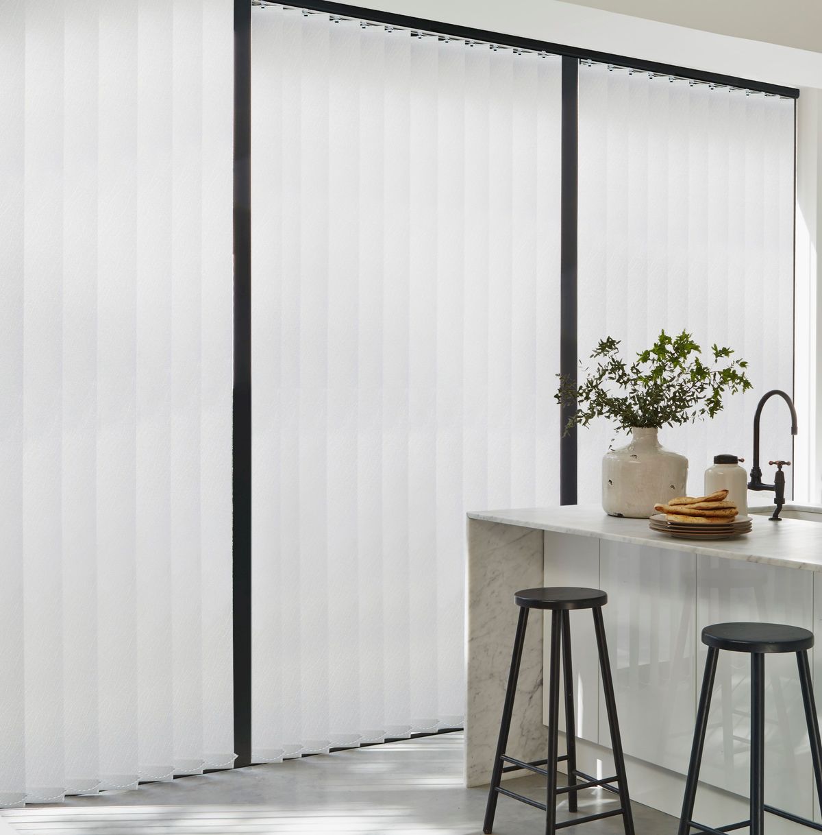 White coloured vertical blinds fitted to a wide floor to ceiling window in a kitchen with black stools and a white breakfast bar