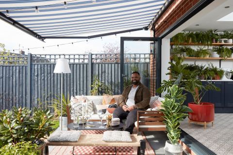 Awnings in a garden hanging over the garden. An influencer sits on an outdoor sofa facing the camera. Surrounded by potted plants. 