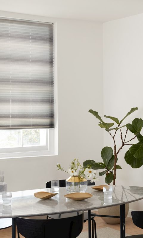 Grey pleated blinds fitted to a window in a dining room with a glass dining table and black chairs