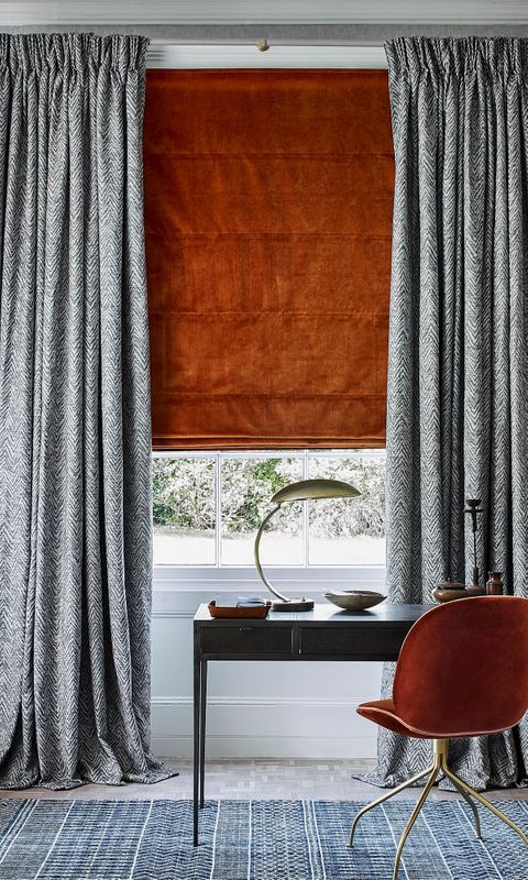 A dark desk and bright orange chair sit in front of a window with a orange blind. The blind is combined with grey detailed curtains. There are also two fabric wall hangings on either side of the window.