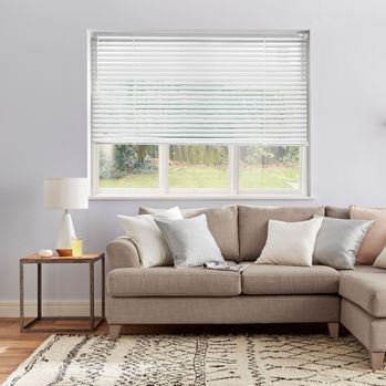 A white coloured metal venetian blind fitted to a rectangular shapes window in  a living room with neutral coloured walls and brown L shaped sofa and sidetable with a lamp