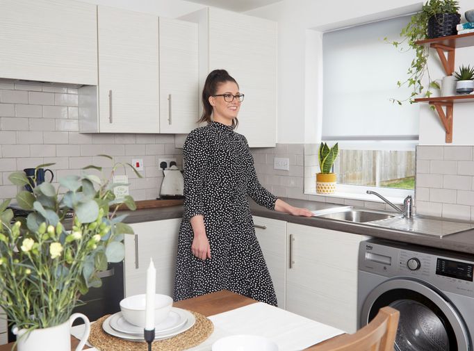 Laura jackson standing in a white and grey decorated kitchen with a white roller blind fitted to a rectangular window. Next to Laura is a wooden dining table with chairs and vase of flowers