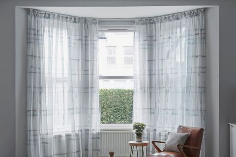 Black striped white voile curtains fitted to a bay window in a living room with brwn leather armchair, wooden coffee table and side table