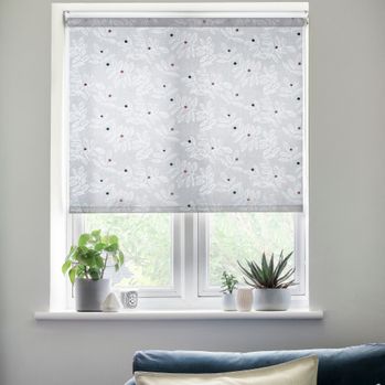 A white coloured Roller Blind with a repeating leaf pattern and multicoloured dots is fitted to a tall rectangular window with potted plants on the window sill, in a living room with a navy blue sofa and white decorated wall