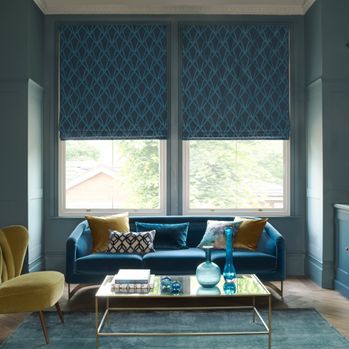 Dark roman blinds at a window in a living room. A sofa faces away from the window and there is a table in the center of the room and a smaller chair on the left side. 