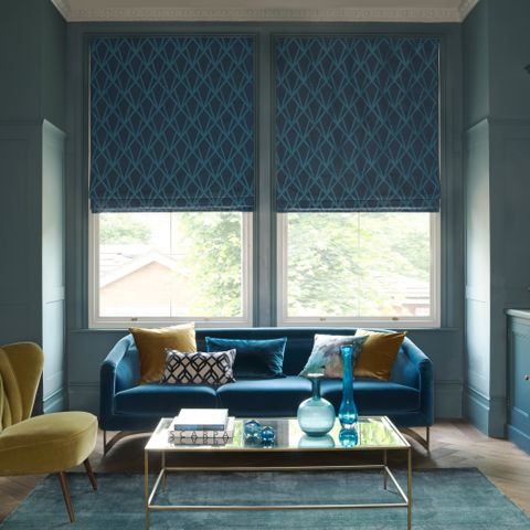 Dark roman blinds at a window in a living room. A sofa faces away from the window and there is a table in the center of the room and a smaller chair on the left side. 