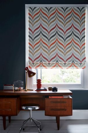 Roman blinds with a dark navy wall and a wooden desk and metal stool. There is a lamp and ornaments on the desk. 