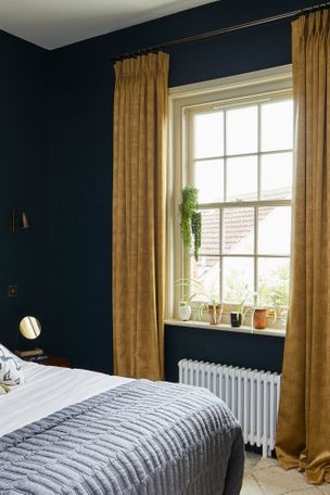 Mustard curtains in a bedroom. There is a bed on the left hand size that takes up the room. There is plotted plants in the room by the window and bed. 
