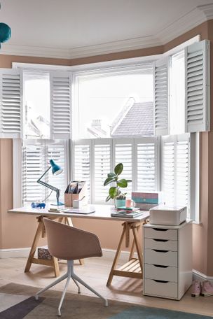 White tier on tier shutters in an office room. There is a desk with office equipment on it and a chair in front of the desk. There is a lamp and a potted plant on the desk.  