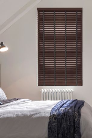 Dark brown faux wood blinds in a bedroom with a white bed and a lamp overhead. 