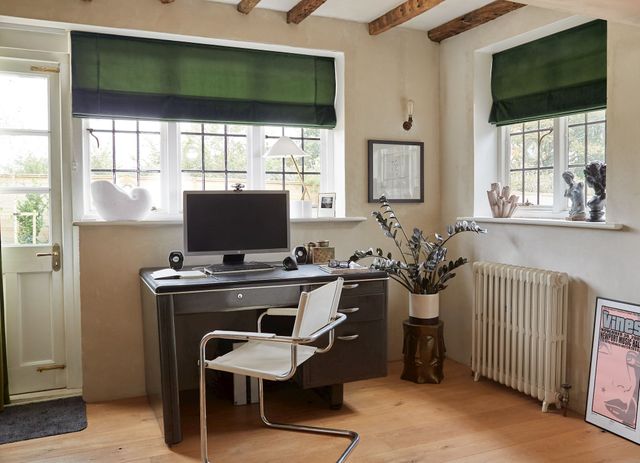 A desk with a computer on it and a chair at the desk with green roman blinds at the window. 