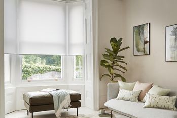 an off white, oyster coloured roller blind on a large window in a tall living room containing a brown stool and light grey sofa