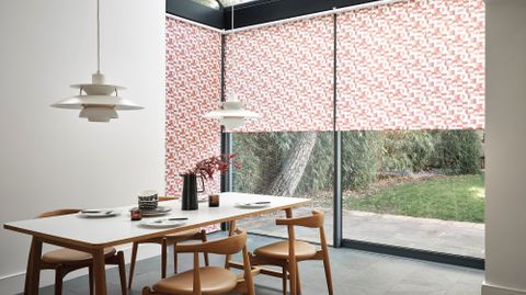 Red and white patterned roller blinds fitted to tall windows in a dining room with dining table and chairs