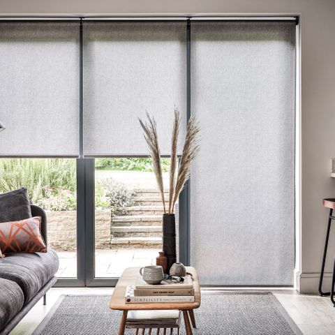 Grey roller blinds in a living room with a grey sofa on the left a table in the centre and a breakfast bar with stools on the right.