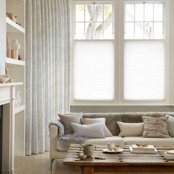 Corner view of a living room where white curtains featuring light grey pattern on curtains dressed over cafe style Pleated blinds on windows.