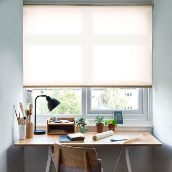 Reber Cream Roller Blind with a desk in front of the window and a lamp on the table and office stationary