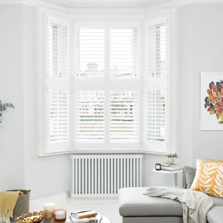 Tier on Tier white shutters in a bay window in a white living room with a radiator under the window. A grey sofa to the right and a painting on the wall above it.